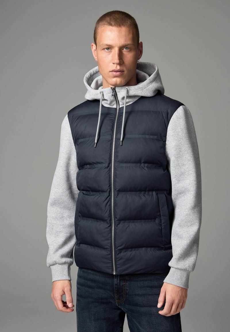 Doudoune marine sans manches avec un hoodie gris à manches longues. Présente un design matelassé, une fermeture éclair et des poignets côtelés gris. Portée sur un jean en denim foncé.