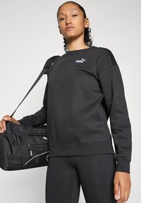 Una donna indossa un felpa nera Puma e leggings, tenendo una borsa da viaggio nera Puma sulla spalla, su uno sfondo semplice.