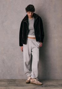 Hnědá fleecová bunda s vysokým límcem, šedé přiléhavé tričko, šedé tepláky a béžové tenisky se světlejšími tkaničkami a texturovanou podrážkou.