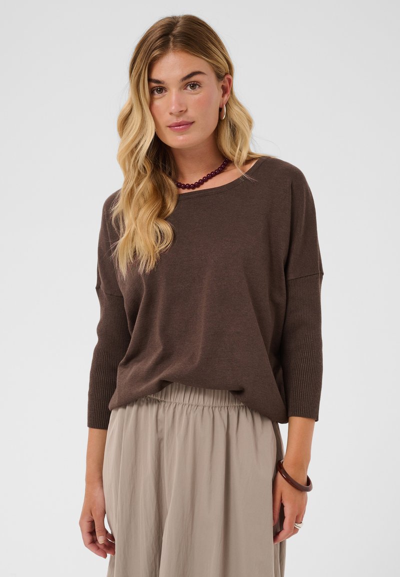 Femme blonde aux ondulations lâches portant un pull marron à manches longues, un pantalon beige à taille élastique, un collier violet en perles et un bracelet marron.