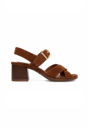 Brązowy zamszowy sandał z odkrytymi palcami, niskim blokowym obcasem, regulowanym paskiem wokół kostki i złotą klamrą, widok z boku na białym tle.