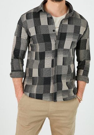 Man draagt een zwart-wit geruit overhemd met opgerolde mouwen over beige broek, handen in de zakken, ketting en armbanden zichtbaar.