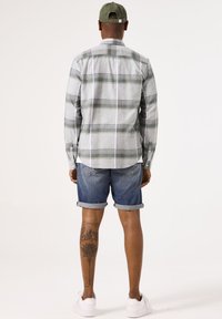 Camisa de franela a cuadros en gris claro y verde, de mangas largas, combinada con pantalones cortos de mezclilla enrollados y zapatillas blancas. Gorra sencilla en la cabeza.