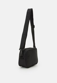 Černá obdélníková krosbody taška vyrobená z hladké tkaniny, s texturovaným popruhem s brandingem, zipovým zapínáním nahoře a kovovými akcenty.