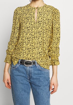 Femme portant un chemisier jaune à manches longues avec un imprimé floral noir, des poignets froncés, associé à un jean bleu et une ceinture noire cloutée.