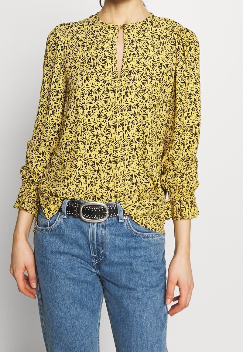 Femme portant un chemisier jaune à manches longues avec un imprimé floral noir, des poignets froncés, associé à un jean bleu et une ceinture noire cloutée.