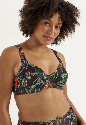 Kvinde med krøllet hår iført en bikini med tropisk bladmønster og matchende nederdel mod en ensfarvet lys baggrund.