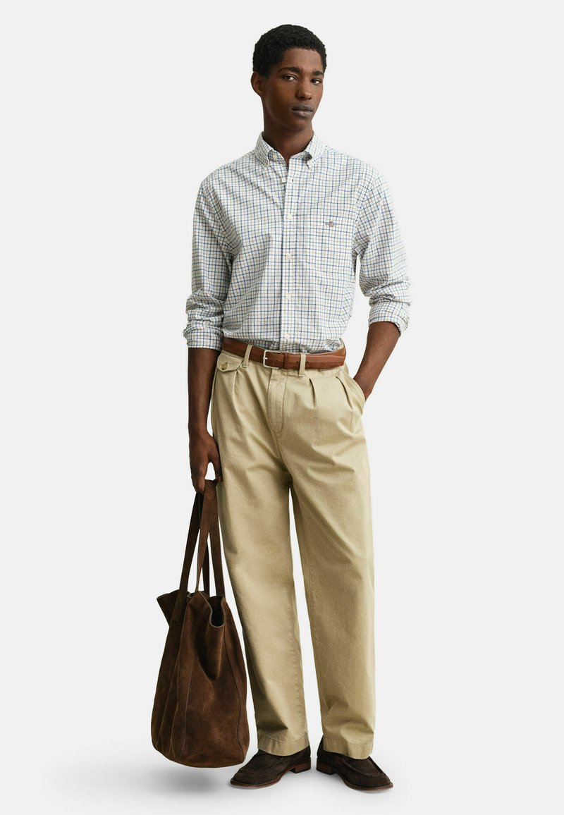 Jeune homme portant un pantalon plissé beige, une chemise à carreaux boutonnée, une ceinture et des chaussures marron, tenant un grand sac fourre-tout en daim marron foncé.