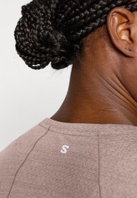 Lysebrun t-skjorte i tekstil som viser bakre nakkeparti med hvit preget logo "S." Har teksturert stoff og buet halslinning.