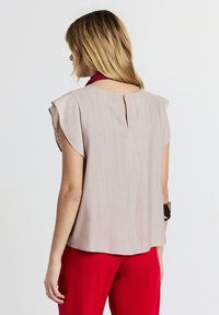 Blusa beige chiaro con maniche a farfalla corte, dettaglio a chiave sul retro e tessuto testurizzato. Abbinata a pantaloni rossi vivaci.