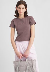 Νεαρή γυναίκα φορώντας μια βυσσινί μπλούζα Nike και ανοιχτό ροζ παντελόνι, κρατώντας μια ανοιχτό γκρι τσάντα tote με το λογότυπο της Nike, στέκεται μπροστά σε έναν απλό φόντο.