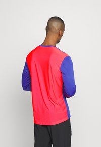 Långärmad skjorta med röd kropp och blå ärmar, tillverkad av ett mjukt, lätt tyg. Rund hals och avslappnad passform.