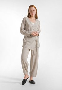 Punatukkainen nainen seisoo päällä, yllään beige kietaisupaita, vastaavat väljät housut, valkoinen aluspaita ja mustat liukukengät yksivärisellä taustalla.