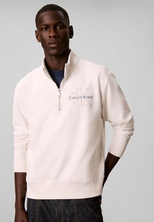 Mann trägt ein weißes Sweatshirt mit halbem Reißverschluss und "Calvin Klein"-Logo, steht mit einer Hand hinter dem Rücken vor einem schlichten grauen Hintergrund.