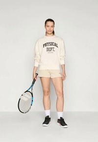 Felpa beige chiaro con la scritta "PHYSICAL DEPT." abbinata a shorts beige. Il modello tiene in mano una racchetta da tennis, indossando scarpe sportive nere e calze bianche.