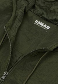 Chaqueta verde, ligera, con textura arrugada, que presenta forro de malla, cremallera frontal y una etiqueta marcada "IUMAN". Talla L.