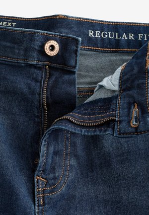 Tamno plave traperice regularnog kroja, s metalnim gumbom, zatvaranjem na patentni zatvarač i kontrastnim narančastim šavovima. Unutrašnjost prikazuje svijetlu tkaninu.