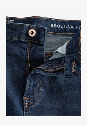 Tamno plave traperice regularnog kroja, s metalnim gumbom, zatvaranjem na patentni zatvarač i kontrastnim narančastim šavovima. Unutrašnjost prikazuje svijetlu tkaninu.