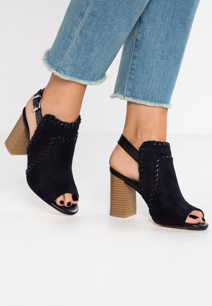 Fødder i sorte åbne højhælede sko med syninger, parret med lyseblå jeans med frynser.