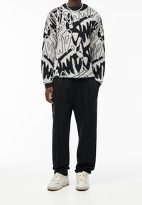 Person wearing a black-and-white abstract patterned sweater, black drawstring pants, and white sneakers against a white background.