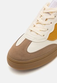 Sneaker, mille kreemjast nahast ülaosast, pruuni seemisnahast aktsentidega ja tekstureeritud kummist tallaga. Omab kreemjaid paelu ja lainekujulist disainidetaili.