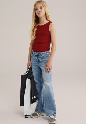 Top senza maniche rosso abbinato a jeans larghi strappati di un blu chiaro. L'outfit include una giacca varsity bianca e nera e sneakers grigie.