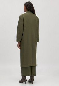 Langer olivgrüner Mantel mit oversized Passform, weiten Ärmeln und minimalistischem Design. Getragen über passenden, cropped Hosen und Absatzstiefeln.