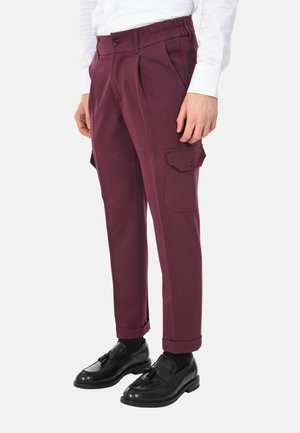 Uomo che indossa pantaloni cargo bordeaux con tasche laterali, mocassini neri con nappine e una camicia bianca, in piedi davanti a uno sfondo semplice.