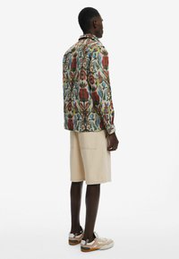 Langärmliges Hemd mit Blumenmuster in lebhaftem Rot, Blau und Grün auf einem cremigen Hintergrund, kombiniert mit beigen Shorts und bunten Turnschuhen.
