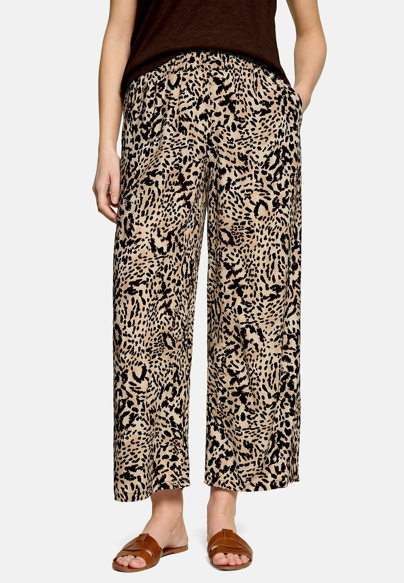Femme portant un pantalon beige à jambes larges avec des taches abstraites noires, un haut marron sans manches et des sandales marron ouvertes, mains dans les poches.