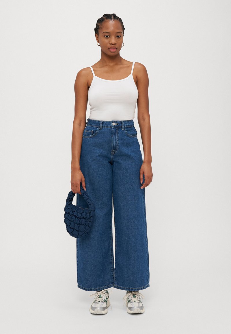Videbukser i blått, hvit tanktop, og en flettet navy håndveske. Sneakers med hvite og grønne detaljer. En enkel bakgrunn.