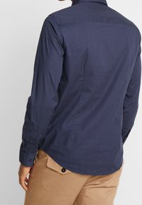 Mörkblå långärmad skjorta med ett subtilt geometriskt mönster, knäppning vid ärmsluten och rundad nederkant, gjord av ett mjukt tyg.