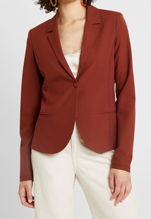 Mujer con blazer ajustado color óxido con cierre de un solo botón, combinado con pantalones crema y grandes pendientes redondos naranjas.