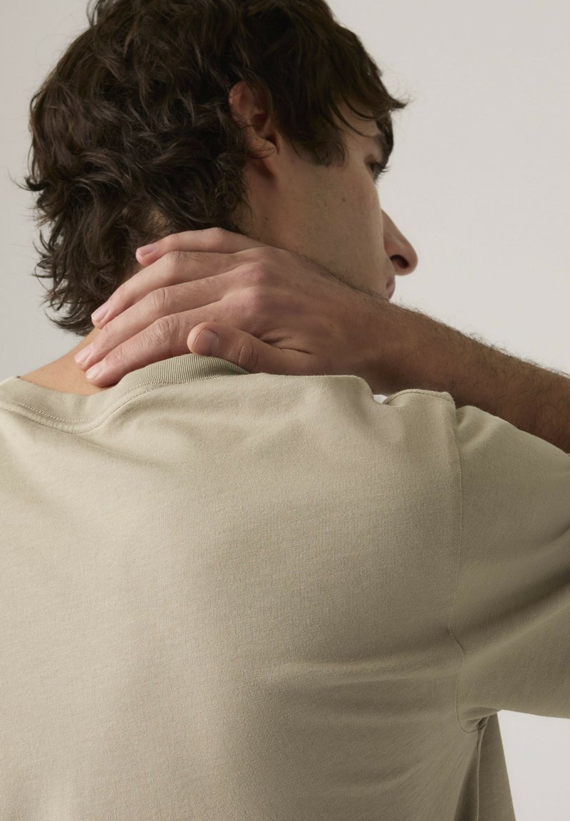 Uomo con capelli castani che indossa una camicia beige, con la mano sinistra che tiene la parte posteriore del collo, su uno sfondo chiaro e uniforme.