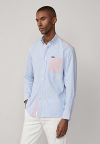 Camicia a maniche lunghe con bottoni, caratterizzata da righe blu e bianche, con una toppa rosa a contrasto sulla spalla sinistra e sulla tasca, realizzata in cotone.