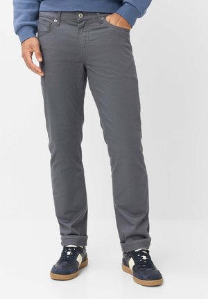 Man wearing dark gray pants, navy blue sneakers with white accents, and a blue long-sleeve top, standing with one hand in pocket.