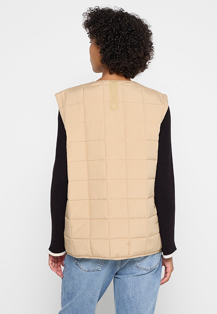 Beige gesteppte Weste mit einem quadratischen Muster, ärmellosem Design und einem hellbraunen Knopfdetail auf der Rückseite, getragen über einem schwarzen Langarmshirt.