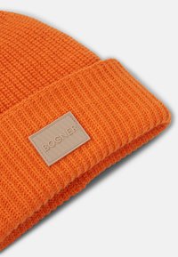 Světlé oranžové pletené čepice s ohrnutým lemem a béžovou obdélníkovou cedulkou s vyraženým nápisem "BOGNER" na lemu.