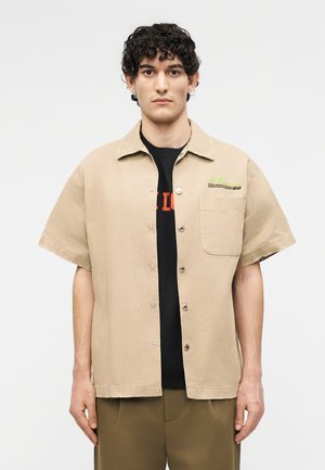 Νέος άνδρας με σγουρά μαλλιά που φοράει μπεζ κοντομάνικο πουκάμισο με κουμπιά πάνω από μαύρο μπλουζάκι με σχέδιο και χακί παντελόνι, στέκεται μπροστά σε ουδέτερο φόντο.