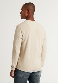 Beige Strickpullover mit langen Ärmeln und strukturiertem Stoff, rundem Ausschnitt und gerippten Bündchen, kombiniert mit dunkelblauen Jeans.