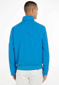 Blaues Bomberjackett mit hohem Kragen, elastischen Bündchen und strukturiertem Stoff. Zeigt Branding am Kragen und ein kleines Logo-Akzent auf der Rückseite.