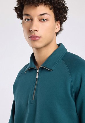 Jeune personne aux cheveux bouclés portant un pull zippé bleu sarcelle, regardant légèrement sur le côté sur un fond clair uni.