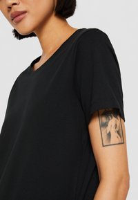 Black cotton t-shirt with short sleeves and a rounded neckline, showing a smooth texture. Visible tattoo on the upper arm.