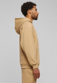 Beige hettegenser laget av mykt stoff, med en stor frontlomme, snor i hetten og ribbestrikkede mansjetter. Enkelt, avslappet design.
