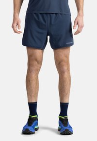 Marina atletiska shorts med en slät yta och elastisk midja. Kombineras med blå och svarta skor med gröna accenter.