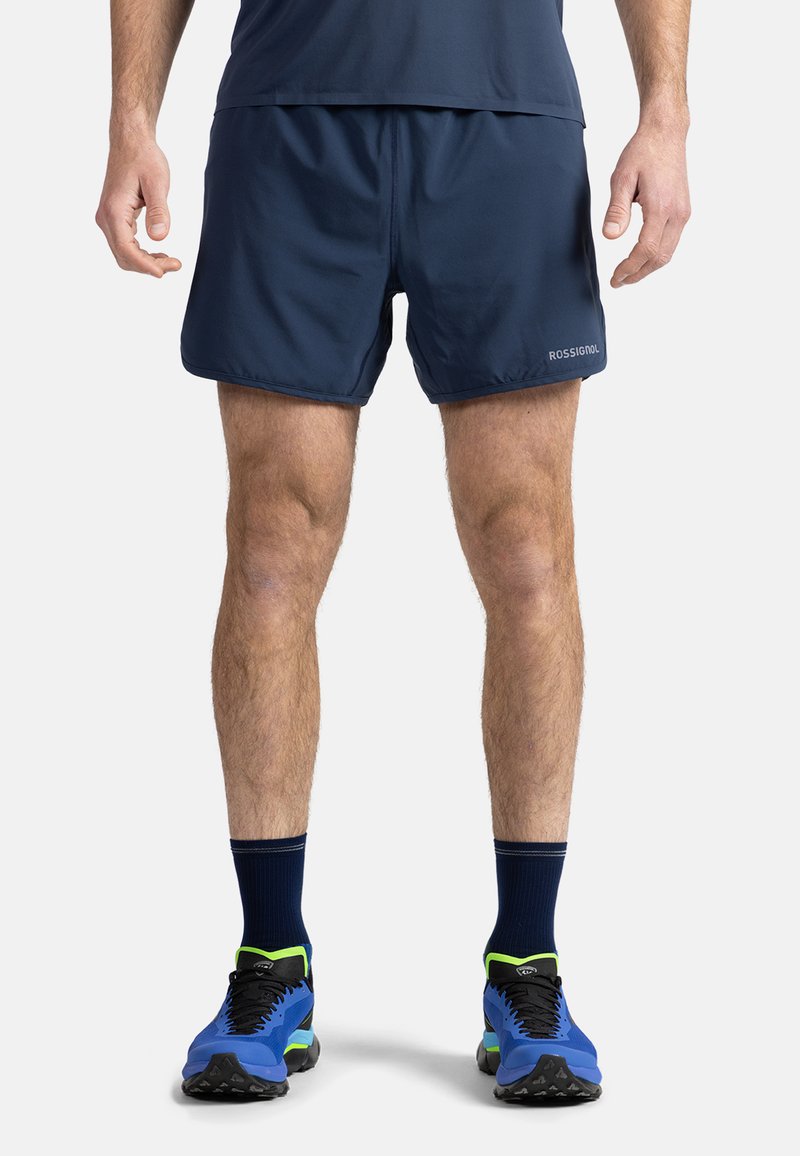 Marina atletiska shorts med en slät yta och elastisk midja. Kombineras med blå och svarta skor med gröna accenter.