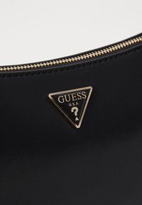 Svart slevitra bag med en gyllen glidelås og en triangelformet gylden logo-plakett med "GUESS" og et spørsmålstegn. Glatt tekstur.