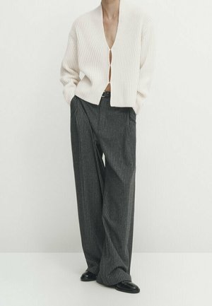Personne portant un cardigan blanc côtelé boutonné au niveau de la poitrine, un pantalon large gris à fines rayures et des chaussures noires, debout les mains dans les poches.