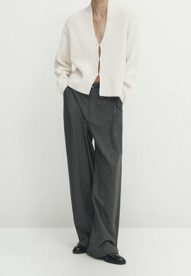 Personne portant un cardigan blanc côtelé boutonné au niveau de la poitrine, un pantalon large gris à fines rayures et des chaussures noires, debout les mains dans les poches.
