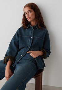 Denim jasje met een losse pasvorm, donkerblauwe kleur, knoopsluiting en contrasterende olijfgroene manchetten. Draagt over bijpassende spijkerbroeken.
